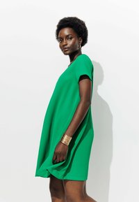 Robe verte à manches courtes avec une coupe ample et fluide. Fabriquée en tissu léger, elle présente un col rond et une texture lisse. Accent avec un bracelet doré.
