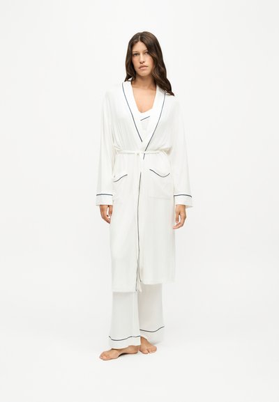 Femme portant une longue robe blanche et un pantalon de pyjama assorti avec un liseré noir, debout pieds nus sur un fond clair.