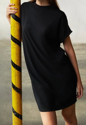 Person, die ein schwarzes kurzärmeliges Kleid trägt und in einem Industriebereich eine gelbe Stange mit schwarzen diagonalen Streifen hält.