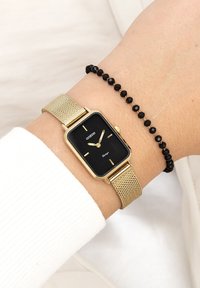 Montre à bracelet en maille dorée avec un cadran rectangulaire noir, présentant des marqueurs et aiguilles dorés, accompagnée d'un bracelet noir à perles sur un poignet.
