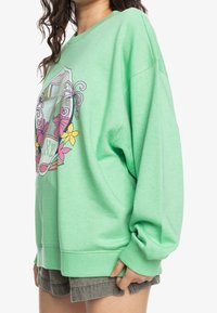 Sudadera extragrande de color verde claro con un diseño gráfico que presenta flores. Textura suave, puños y dobladillo de canalé. Ajuste casual y cómodo.
