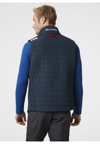 Granatowa ocieplana kamizelka o pikowanej fakturze, z wysokim kołnierzem i czerwonymi akcentami z tyłu, noszona na niebieskiej koszuli z długim rękawem.