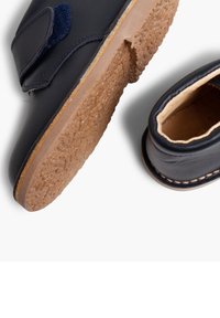 Zapatos de cuero en azul marino con una suela de goma marrón texturizada. Presentan una correa de Velcro y un borde cosido alrededor de la abertura.