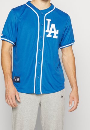Hombre con camiseta azul de béisbol de Los Angeles Dodgers con ribetes blancos y pantalones deportivos grises, de pie frente a un fondo liso.