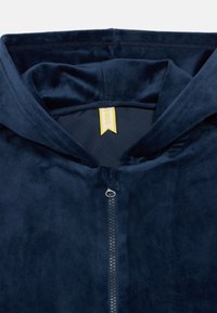 Tumedat sinise värvi kapuutsiga fliis, millel on pehme tekstuur, esiosa tõmblukk ja stringidega kapuuts. Kollane silt on nähtav kaelaaugu seest.