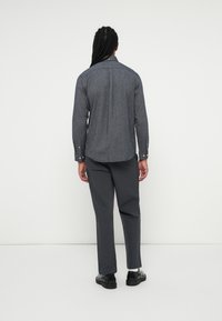 Persoană cu păr lung împletit, purtând un tricou alb cu mânecă lungă, pantaloni gri închis, șosete albe și pantofi negri, stând în fața unui fundal alb.
