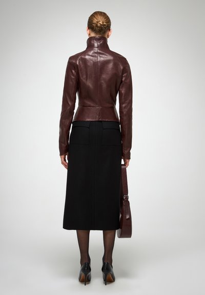 Veste en cuir marron avec col montant, design ajusté et manches longues, associée à une jupe noire longueur genou et des talons noirs, tenant un sac marron.