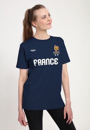 Kobieta z kucykiem ubrana w granatową koszulkę piłkarską Francji z emblematem koguta, napisem "FRANCE" oraz dwoma gwiazdkami z przodu.