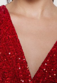 Nahaufnahme einer Person mit freiem Oberkörper und Hals, die ein rotes, mit Pailletten besetztes V-Ausschnitt-Oberteil trägt.