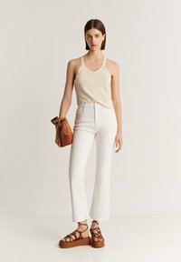 Un débardeur en tricot beige, un jean blanc évasé taille haute et des sandales à plateforme marron. Le modèle tient un sac à main multicolore avec un cordon de serrage.