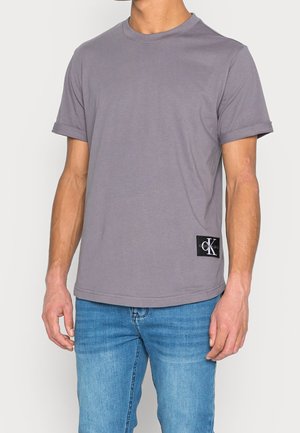 Mężczyzna w szarej koszulce z podwiniętymi rękawami z czarną naszywką logo Calvin Klein i niebieskich dżinsach, stoi na jednolitym tle.