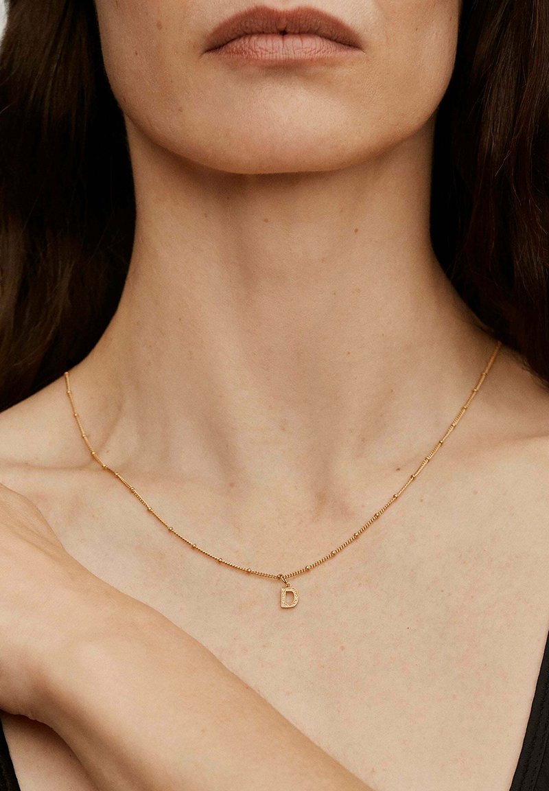 Collar dorado con una delicada cadena y un pequeño colgante de la letra "D" que brilla. La cadena cuenta con cuentas redondas y segmentos.