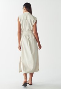Donna che indossa un vestito midi senza maniche beige con cintura e sandali neri con tacco, in piedi contro uno sfondo bianco, di spalle.