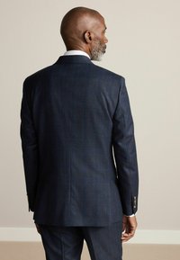 Veste de costume à carreaux marine avec une coupe droite, deux boutons et un soufflet simple ; présente des accents en bouton marron et une texture de tissu lisse.