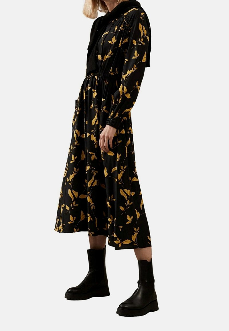 Femme portant une robe midi noire avec des motifs de feuilles jaunes, un pull noir drapé sur les épaules et des bottines noires épaisses.