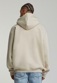 Felpa beige con un cappuccio ampio e strutturato, vestibilità over size e polsini a coste; abbinata a jeans chiari strappati.