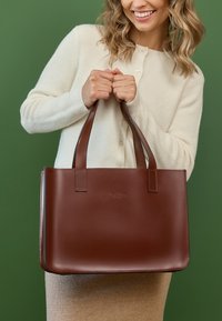 Borsa a tracolla in pelle marrone con due maniglie, superficie liscia e un logo impresso sottile. Posizionata su uno sfondo verde.