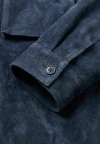 Mørkeblå semsket jakkeerm med én svart knapp ved mansjetten, som viser myk tekstur og fine sømdetaljer.