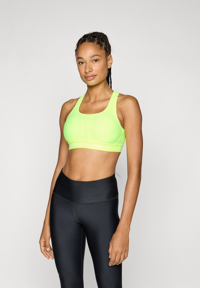Neon grønn sportsbh med brede stropper, logobånd og glatt tekstur, paret med høyhælte svarte leggings.