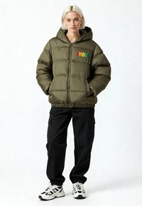 Doudoune verte olive avec capuche, ornée d'une broderie multicolore "AMOUR", fermeture éclair et poignets côtelés. Associée à un pantalon cargo noir et des baskets.