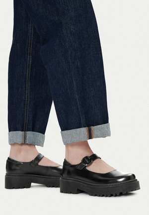 Osoba nosící tmavé zkroucené džíny a černé platformové boty Mary Jane s tlustou podrážkou a páskem na přezku.