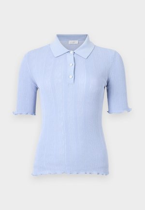 Vrouw met donker haar die een lichtblauw kortemouwenhemd met knopen draagt, donkere wijde spijkerbroek en zwarte hakken, staande tegen een effen witte achtergrond.