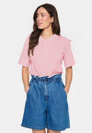 Rosa Polohemd mit kurzen Ärmeln und Kragen, kombiniert mit hochgeschnittenen blauen Jeansshorts, die einen Gürtel und Fronttaschen aufweisen.