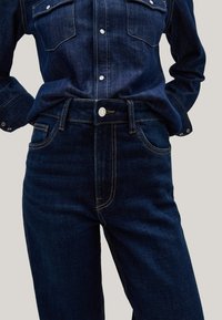Jeans de denim azul oscuro y camisa de denim a juego con botones de presión blancos, llevados por una persona con las manos en los bolsillos frente a un fondo claro.