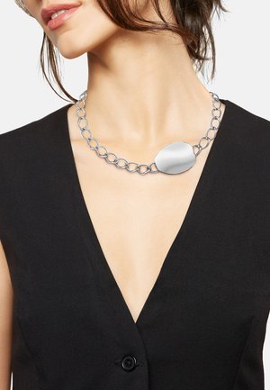 Srebrny naszyjnik z łańcuszka z dużymi, gładkimi owalnymi ogniwami oraz wypolerowanym owalnym wisiorkiem. Noszony z głębokim dekoltem w kształcie litery V w czarnym stroju.