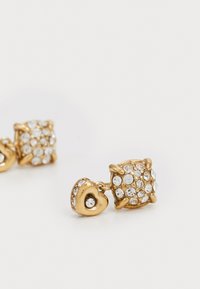 Gouden oorbellen met een hartvormig en een kubusvormig ontwerp, versierd met meerdere kleine heldere kristallen die zijn gezet in een gestructureerde metalen behuizing.