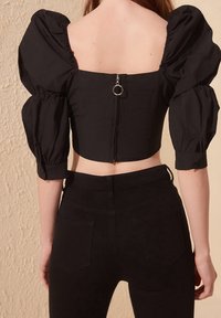 Blusa nera accorciata con maniche a palloncino, scollatura quadrata e cerniera sul retro con tirante in metallo. Abbinata a jeans neri aderenti.