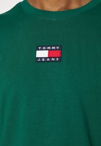 Grön bomull T-shirt med en marinblå tygflicka med texten "TOMMY JEANS", röda och vita detaljer, samt rundad halsringning.