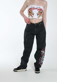 Jeans baggy de mezclilla negra con bordados coloridos de calaveras y flores en la pierna izquierda, combinados con una blusa blanca sin tirantes con gráficos de tigres.