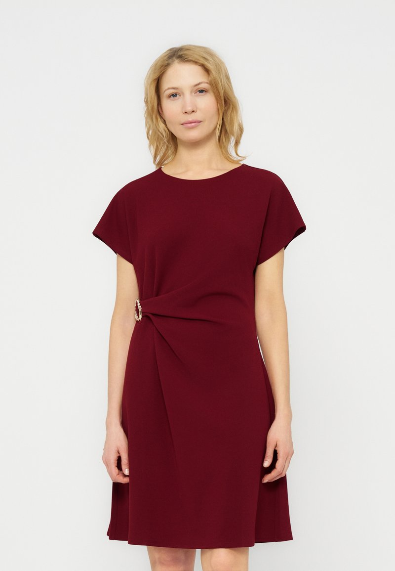 Γυναίκα που φοράει ένα μπορντό φόρεμα με κοντά μανίκια και μήκος μέχρι το γόνατο, με διακοσμητική αγκράφα στο πλάι, στέκεται μπροστά σε ένα απλό λευκό φόντο.