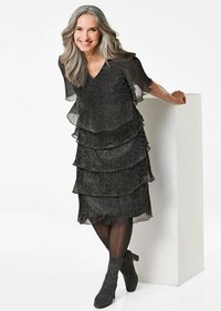 Robe noire à plusieurs niveaux avec des manches volantées en tissu transparent et texturé. Assortie à des bottines noires et à des collants transparents.