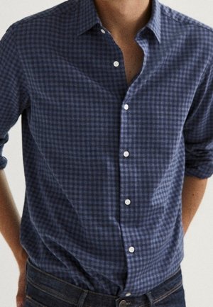 Mand iført en blå og sort ternet skjorte med knapper, ærmerne rullet op, og mørkeblå jeans.