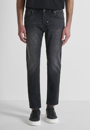 Uomo che indossa jeans neri sbiaditi e sneakers nere con suola bianca, in piedi davanti a uno sfondo grigio chiaro uniforme.