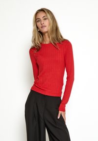 Suéter rojo ajustado de manga larga con textura acanalada y pequeñas perforaciones. Combinado con pantalones negros de tiro alto. Diseño simple y clásico.