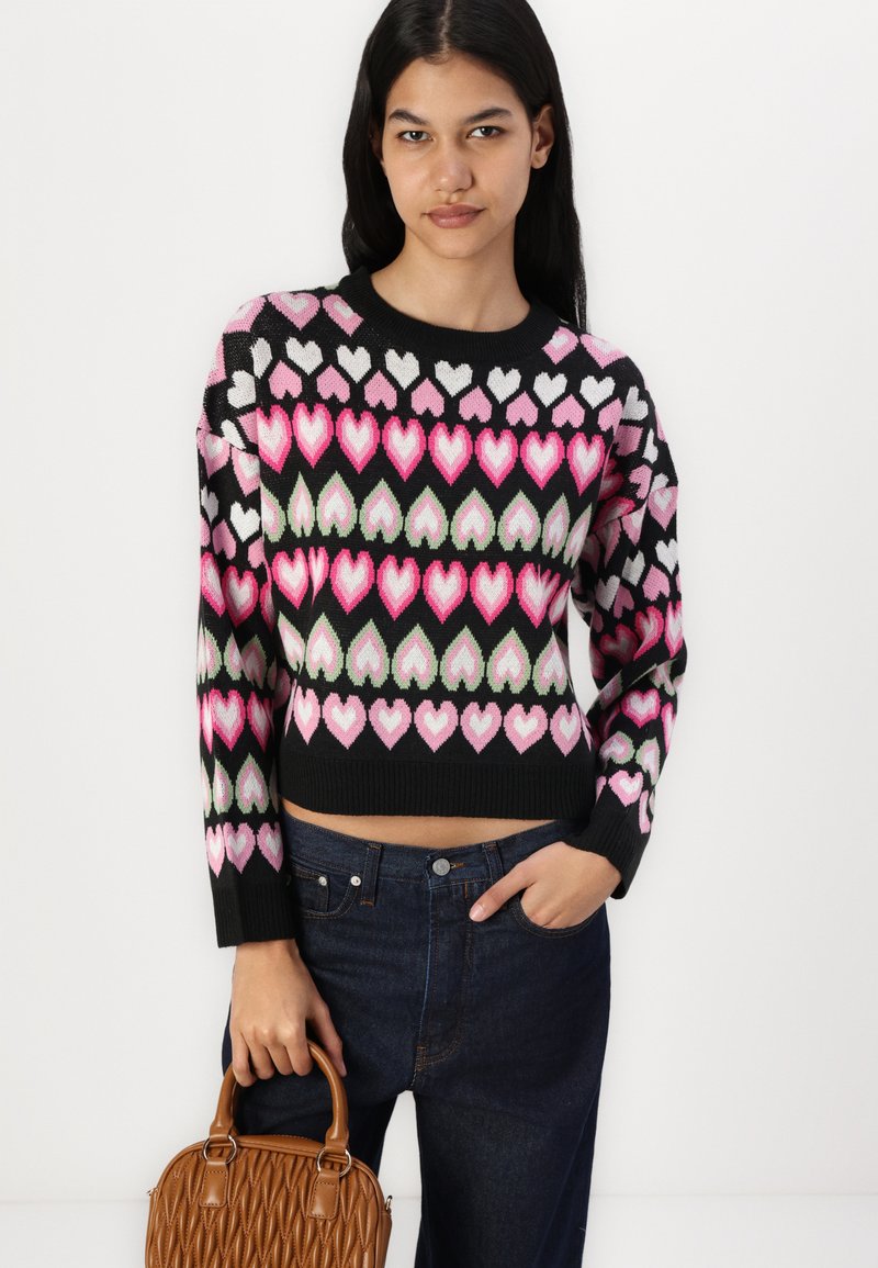 Maglione corto nero con motivo a cuore in rosa, bianco e verde; indossato con jeans a vita alta blu scuro e una borsa marrone.
