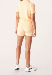 Žena oblečená v lehce žlutých šortkách a odpovídajícím tričku, stojící zády k fotoaparátu, doplněná bílými teniskami Nike Air Max.