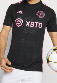 Musta jalkapallopaita, jossa vaaleanpunaiset yksityiskohdat, teksturoidulla kuviolla, Adidas-logo, joukkueen vaakuna ja "XBTO"-logo suurina vaaleanpunaisina kirjaimina.