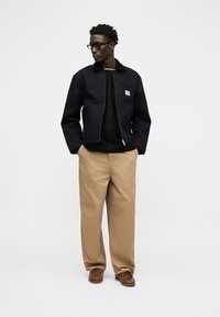 Moški v črni jopiči z zadrgo, črni majici, bež širokih hlačah, rjavih čevljih in očalih stoji z rokami v žepih in gleda levo.