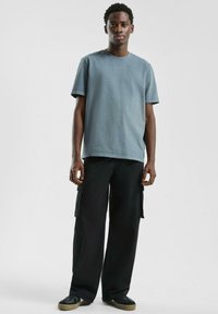 Jonge man die staat in een vervaagde blauwe T-shirt, zwarte cargo-broek en zwarte sneakers met beige zolen, tegen een effen lichte achtergrond.