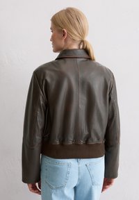 Bruine leren bomberjack met een geribde tailleband, een getailleerde kraag en subtiele stikdetails. Draagt lichtblauwe spijkerbroek.