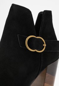 Bota de tobillo de ante negro con punta redondeada, detalle de hebilla lateral en oro y tacón de bloque de madera. La textura parece suave y lisa.