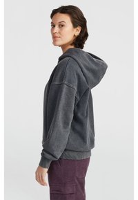 Felpa grigia con cappuccio, dal taglio morbido, con polsini e orlo ribattuti. Il tessuto ha una finitura strutturata. Indossata con pantaloni in velluto a coste viola.