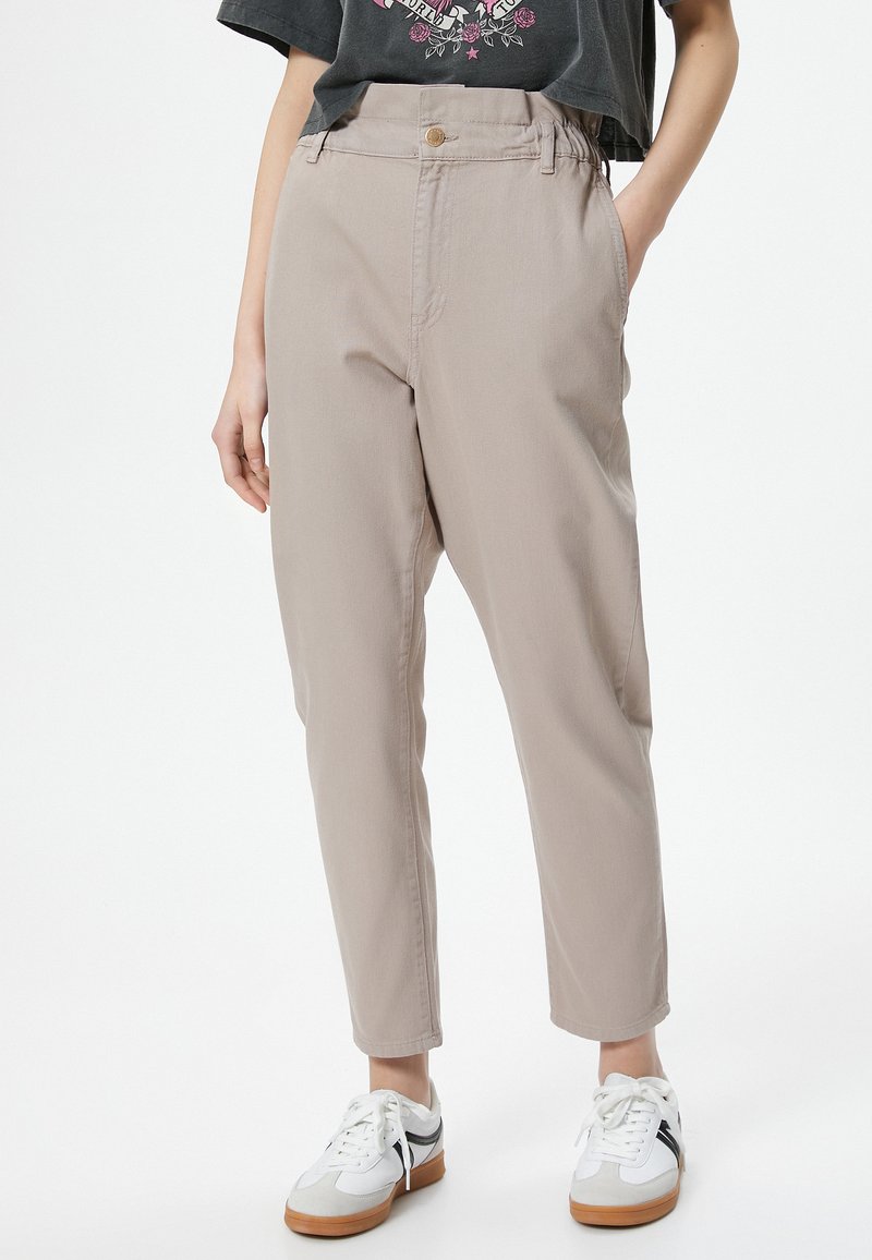 Beige tapered pants with a high waist, which have a front button and pockets. They are paired with white sneakers that have black accents.