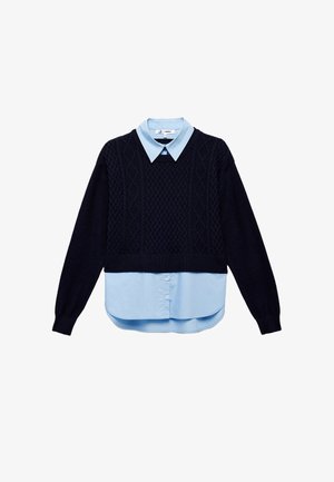 Merinotakki tummansinisestä neuloksesta, jossa on teksturoitu timanttikuvio, kerroksittain vaaleansinisen kauluspaidan päällä. Ominaisuuksiin kuuluu lyhyt malli ja napitus etupuolella.