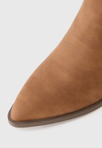 Punta affusolata di un stivale alla caviglia in suede tan con suola marrone scuro su uno sfondo bianco.