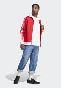 Red athletic jacket with white stripes, white t-shirt, blue denim jeans, black sneakers, and white socks with a red logo, standing on a light background.